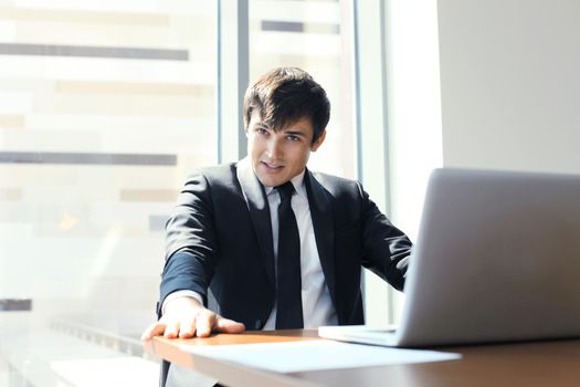 Portrait of happy businessman sitting at office desk, looking at camera, smiling.