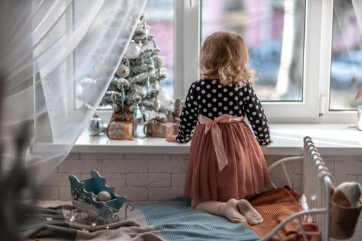 A little girl is sitting on the bed by the window and decorating a small tree with tiny Christmas toys. Happy healthy child celebrating a traditional family holiday. Adorable baby