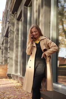 caucasian smiling woman in beige trench coat calling by smartphone outdoors on sunny day. attractive stylish female street portrait at fall or spring. autumn lifestyle, modern communication technology