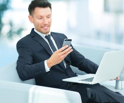 closeup of a businessman sitting in a chair with your smartphone and working on laptop in the lobby of the Bank.the photo has a empty space for your text