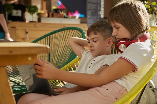 boy and girl sit outdoors together in one chair, watch at screen of laptop on sunny day. little caucasian schoolchildren sitting at veranda of cafe, studying with computer. online distant learning