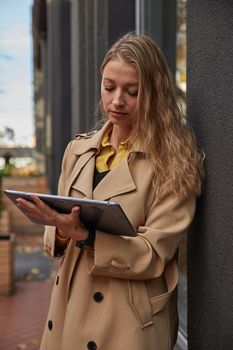 young caucasian woman in coat using tablet outdoors on sunny day, reading ebook, watching movie. attractive female street portrait at fall or spring. autumn lifestyle, modern communication technology