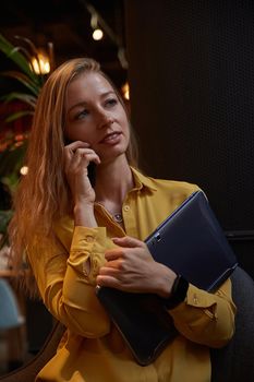 attractive cauasian woman calling by smartphone, holding laptop in hands. beautiful stylish lady talking by phone at cafe. modern communication technology, distant work, remote job, freelance