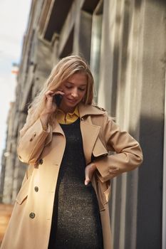 caucasian smiling woman in beige trench coat calling by smartphone outdoors on sunny day. attractive stylish female street portrait at fall or spring. autumn lifestyle, modern communication technology