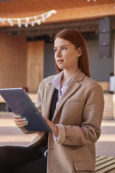 young caucasian woman in coat using tablet outdoors on sunny day, reading ebook, watching movie. attractive female street portrait at fall or spring. autumn lifestyle, modern communication technology