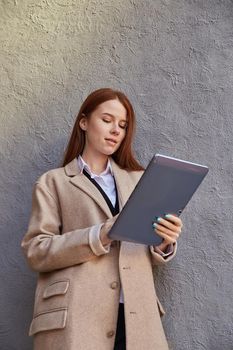 young caucasian woman in coat using tablet outdoors on sunny day, reading news, social media. attractive female street portrait at fall or spring. autumn lifestyle, modern communication technology