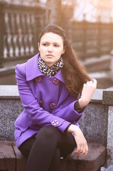 Beautiful girl sitting on a bench in fialetovom coat in clear time of the day, wondering and looking in the direction of hair straightening