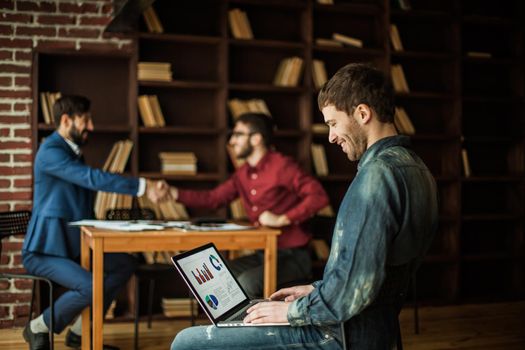 sales Manager working on laptop with a marketing report on the background of business partners handshaking