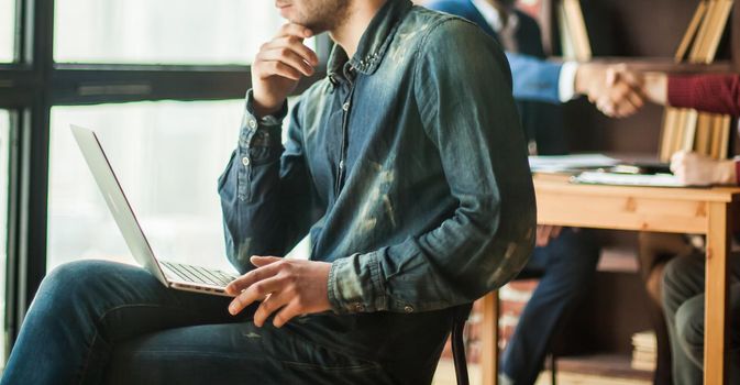 employee of the company with laptop looking out the window on the background of business partners handshaking in the workplace in the office