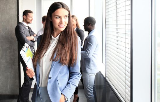 Business woman standing in foreground with a tablet in her hands, her co-workers discussing business matters in the background