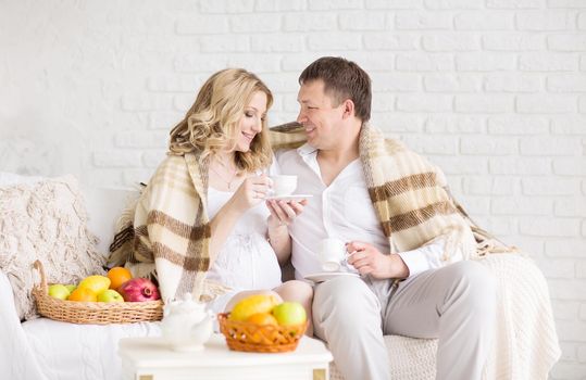 Portrait of a beautiful young couple, pregnant girl having breakfast home having covered plaid