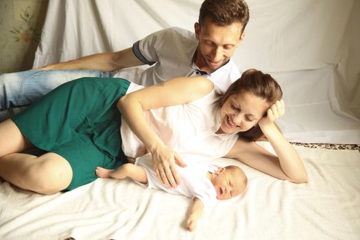 family portrait - happy mother and father lie side by side with a newborn baby on the bed