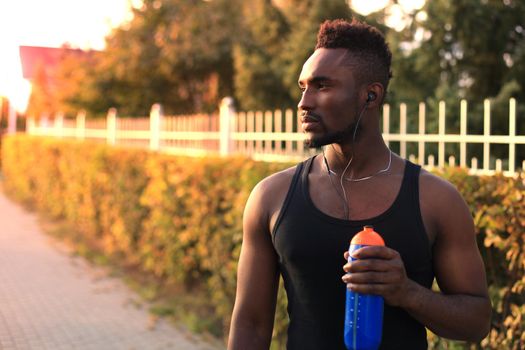 African man in sports clothing drinking water while standing outside, at sunset or sunrise. Runner.