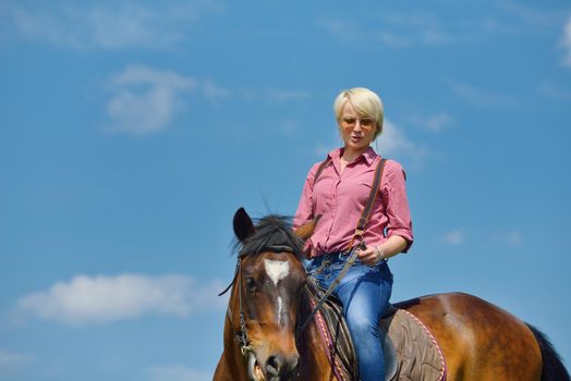 happy woman in sunglasses sitting on horse farm animal outdoors with blue sky in background