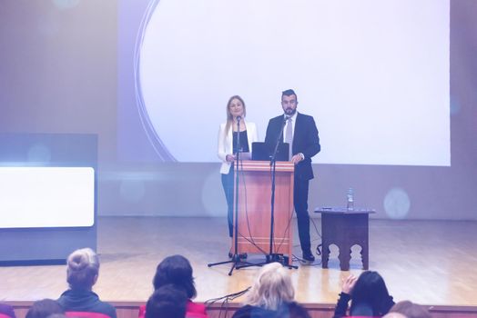 two young businesspeople giving a presentation to a group of colleagues seated in chairs at conference room