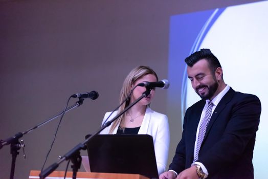 two young businesspeople giving a presentation to a group of colleagues seated in chairs at conference room