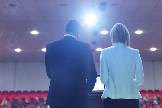 two young businesspeople giving a presentation to a group of colleagues seated in chairs at conference room