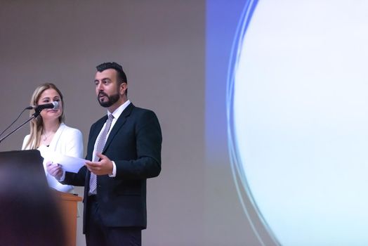 two young businesspeople giving a presentation to a group of colleagues seated in chairs at conference room