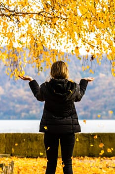 Back view of alone woman enjoying autumn, throwing fallen leaves on autumn alley. Autumn landscape, orange foliage in a park in Orsova, Romania, 2020