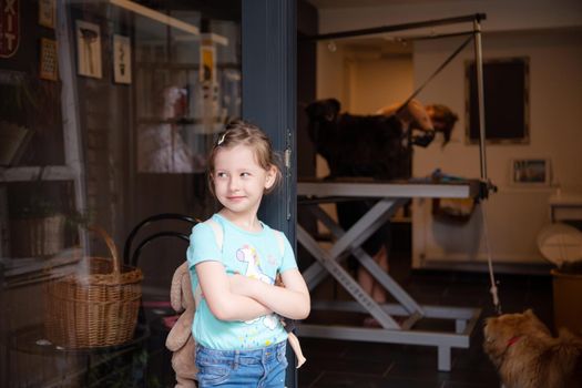 portrait of cute little girl standing and waiting while professional pet hairdresser finishing her black dog hairstyle in front of beauty salon for animals