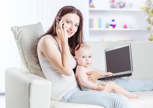 mother holding one year old baby in his arms,talking on his cell phone and working on laptop sitting on sofa in living room