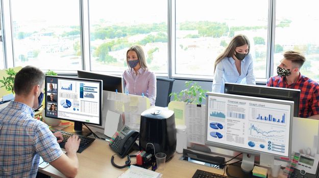 Startup business team in protective masks on meeting in modern bright office interior brainstorming, working on computers