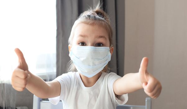 Little girl in a medical mask showing thumb's up gesture. Protection against coronavirus