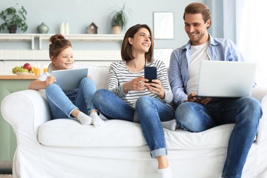 Father, mother and daughter using electronic devices sitting on sofa at living room