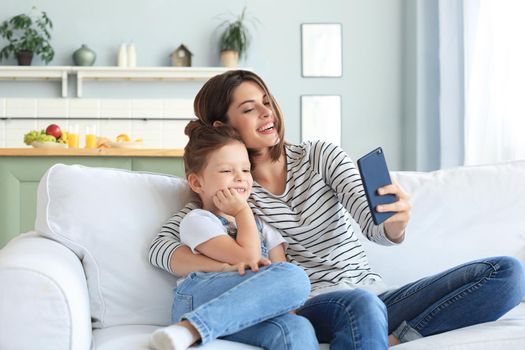 Beautiful young mother and her little daughter taking a selfie with a smartphone at home on the couch