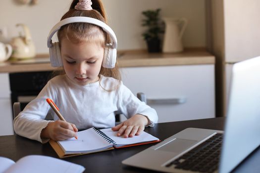 Little girl in headphones sit at desk writing in notebook studying online do exercises at home, little child handwrite prepare homework on quarantine, have web class or lesson indoors