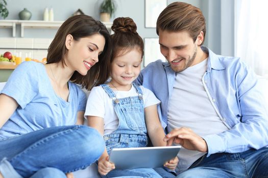 Positive friendly young parents with smiling little daughter sitting on sofa together answering video call on digital tablet while relaxing at home on weekend