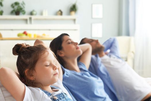Relaxed young family resting and dreaming about new home on comfortable sofa together at home, happy young parents with little daughter relax enjoying nap relaxing or meditating