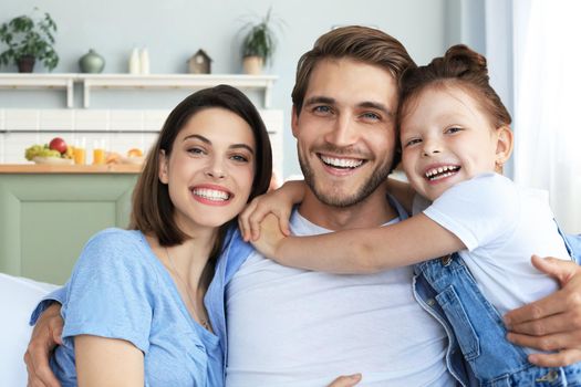 Positive friendly young parents with smiling little daughter sitting on sofa together while relaxing at home on weekend