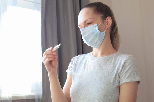 Young woman in the medical mask looks at the thermometer with high temperature