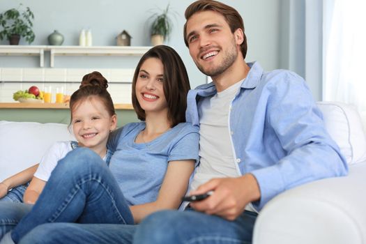 Happy family with child sitting on sofa watching tv, young parents embracing daughter relaxing on couch together