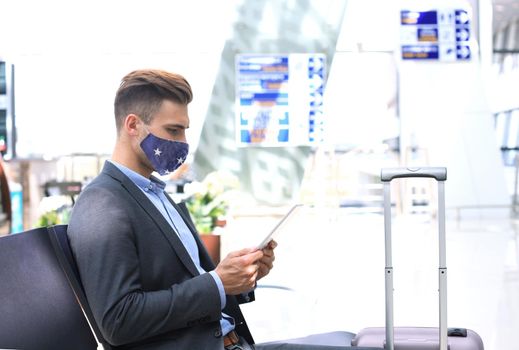 Businessman in protective mask with suitcase using digital tablet in hall of airport. Airport in the coronavirus epidemic.