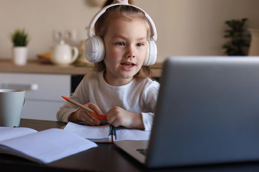 Smiling little girl in headphones handwrite study online using laptop at home, cute happy small child in earphones take Internet web lesson or class on PC