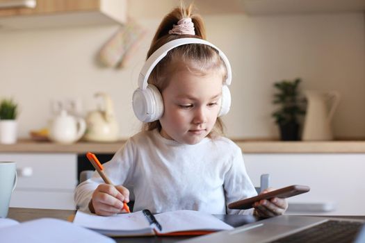 Smart little girl in headphones do homework online class on smartphone, small child in earphones study on Internet, have web conference or lesson during quarantine