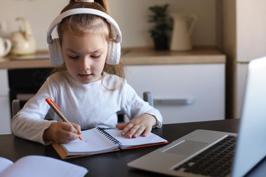 Little girl in headphones sit at desk writing in notebook studying online do exercises at home, little child handwrite prepare homework on quarantine, have web class or lesson indoors