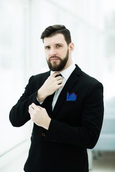 portrait of a successful businessman adjusting his necktie while standing near a window in bright office.the photo has a empty space for your text