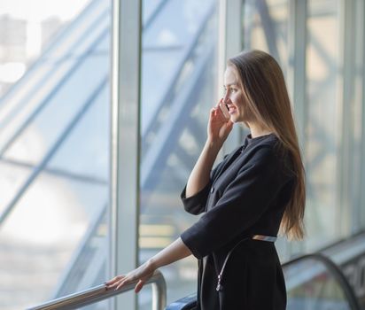 Confident successful business woman with a smartphone, standing in front of a window in a modern office.