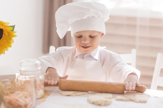 A small child in the form of a cook rolls out the dough on the kitchen table
