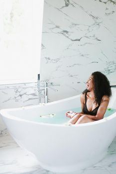 Young happy black woman sitting in bathroom with marble tile, wearing black swimsuit. Concept of personal care and spa.