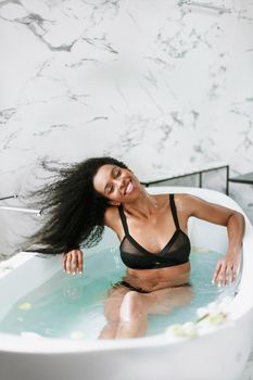 Young cute black woman taking bath in bathroom with marble tile, wearing black swimsuit. Concept of personal care and spa.