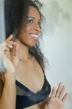 Portrait of young afro american smiling nice young woman wearing bra and touching glass in bathroom. Concept of underwear photo session.