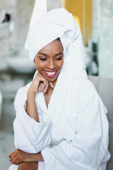 Young cute afro american woman sitting in bathroom and wearing white bathrobe. Concept of relax and personal care.