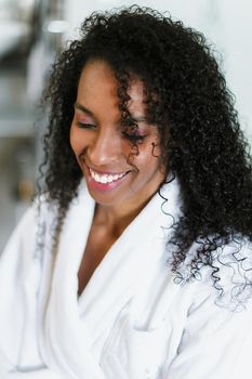 Close up photo of pretty afro american girl sitting in bathroom and wearing white bathrobe. Concept of black beauty and relax