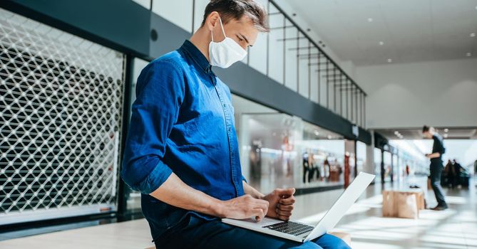 man in a protective mask works on a laptop in a shopping center building. pandemic in the city