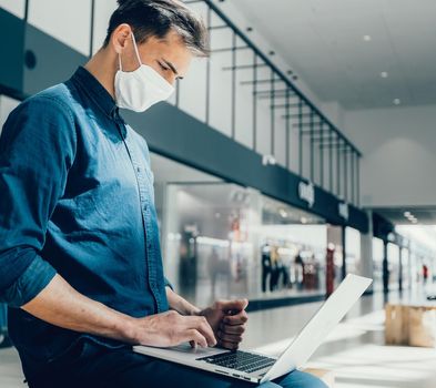 man in a protective mask works on a laptop in a shopping center building. pandemic in the city