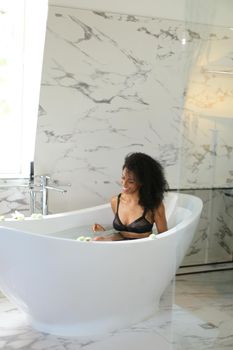 Afro american yougn woman relaxing in bath and wearing bra with panties. Concept of bathroom underwear photo session and morning relax.